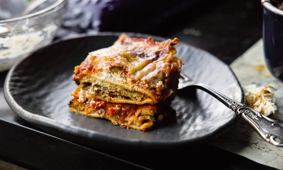 A close-up of a piece of vegan lasagna on a dark plate, with layers of pasta, rich tomato sauce, and melted cheese. A fork rests beside it. The setting has a rustic and cozy feel.